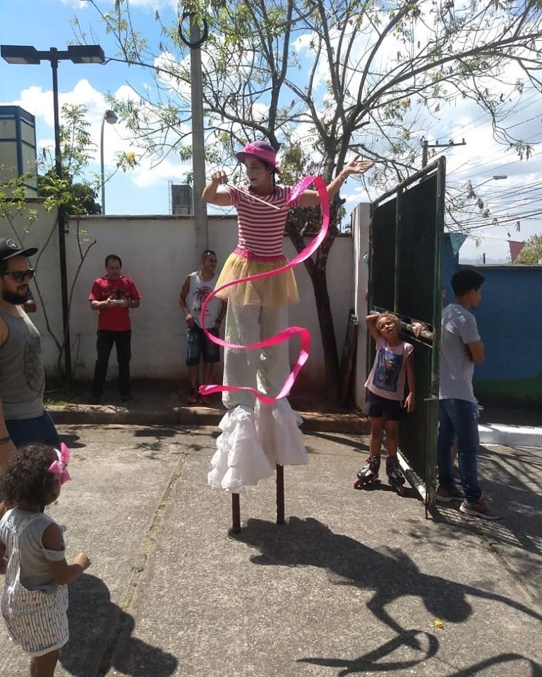 Uma moça, vestida de forma caricata e com rosto pintado, sorri para as crianças que a assistem. Ela está sobre pernas de pau e balança uma fita de cetim Pink. 